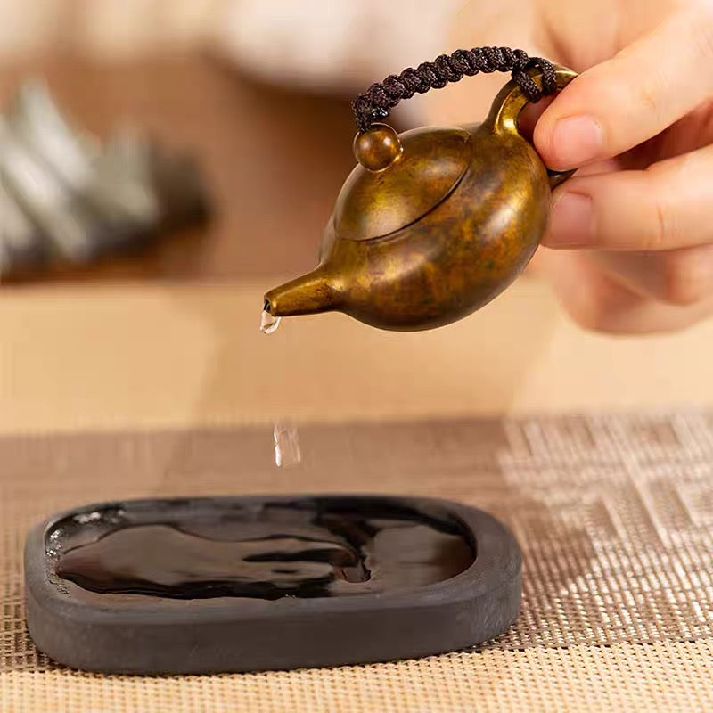 Bronze teapot being used to pour liquid onto a black stone coaster on a woven mat.
